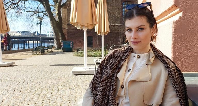 Linda Loran sitzt draußen an einem Tisch am Flussufer, im Hintergrund Sonnenschirme und eine Brücke